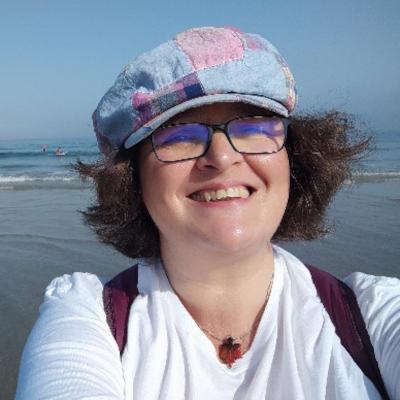 Una donna sulla quarantina con un cappello, occhiali in riva al mare. Ha i capelli tagliati a media lunghezza, paffuta e sorridente.