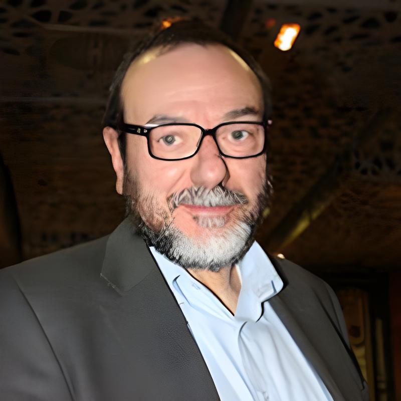 Un uomo stempiato sulla cinquantina con occhiali barba e baffi. Veste elegante.